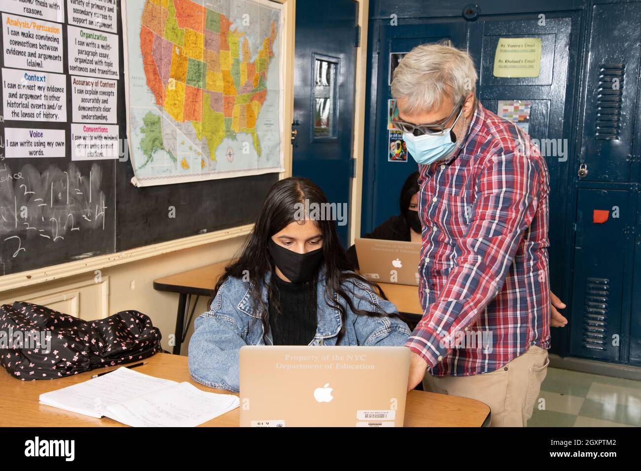 Education High School classroom scene male teacher working with female student in classroom, both wearing face masks to protect vs Covid-19 Stock Photo