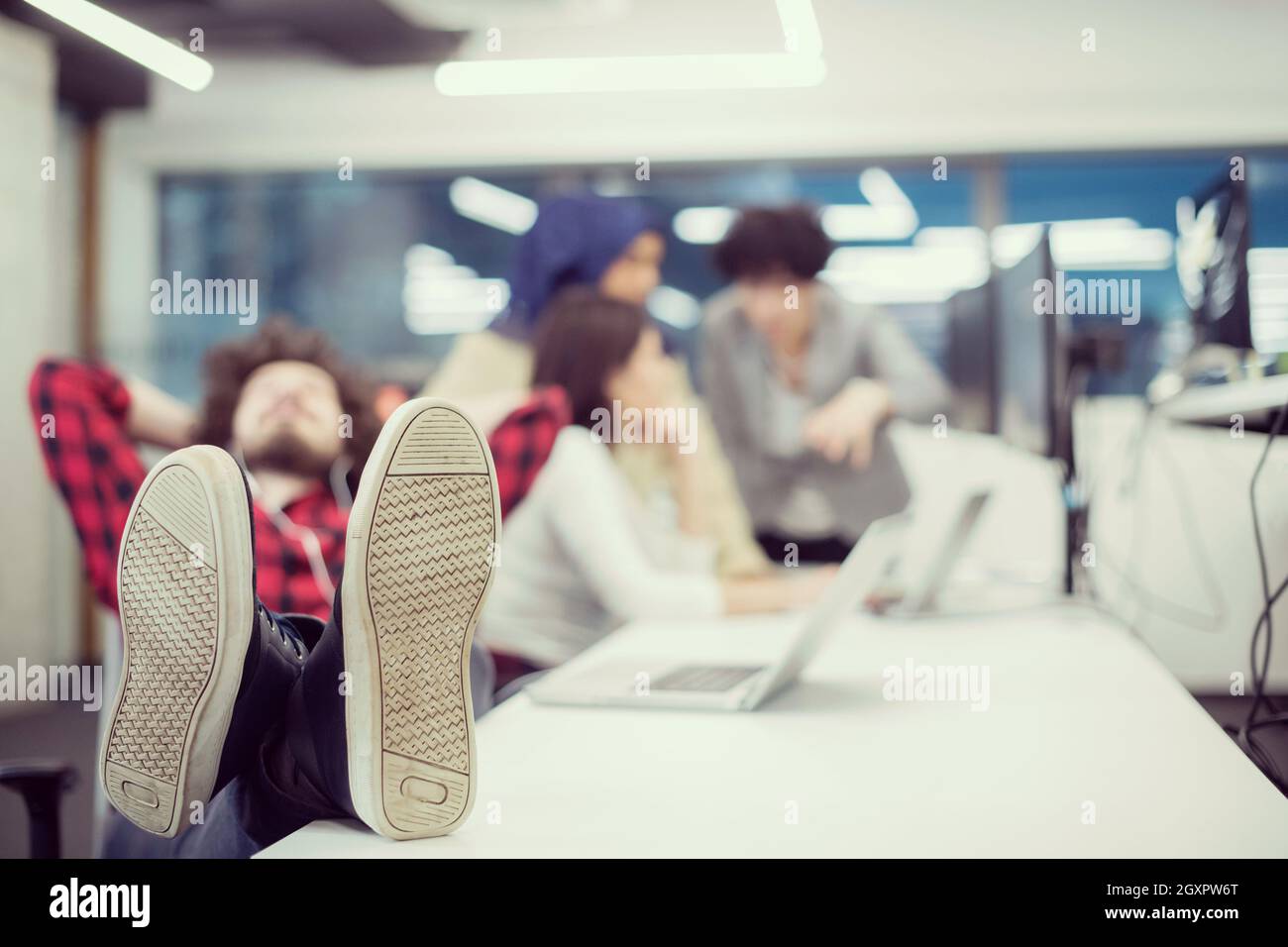 young software developer resting with legs on desk while his multiethnics business team writing programming code in the background at startup office Stock Photo