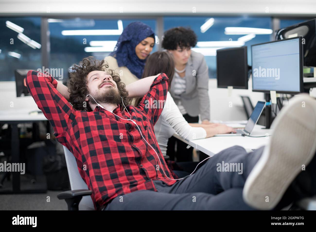 young software developer resting with legs on desk while his multiethnics business team writing programming code in the background at startup office Stock Photo