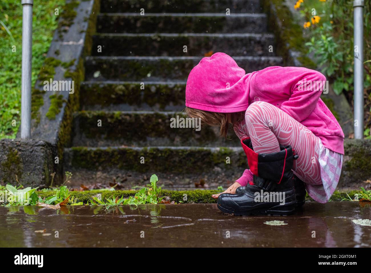 A young child plays on a sidewalk in the rain while wearing pink. Stock Photo