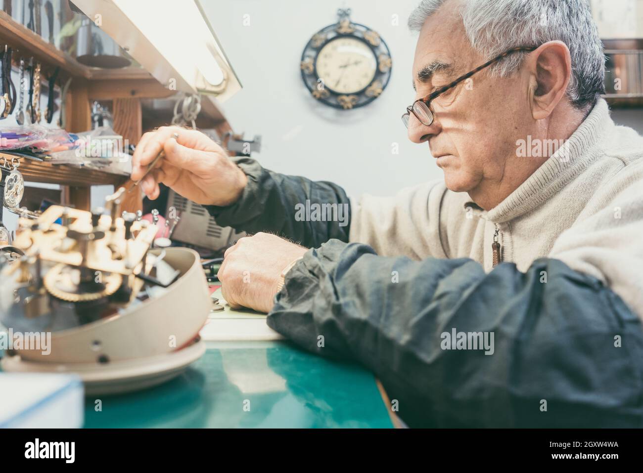 Watchmaker in his workshop repairing a larger clock with clockwork in foreground Stock Photo