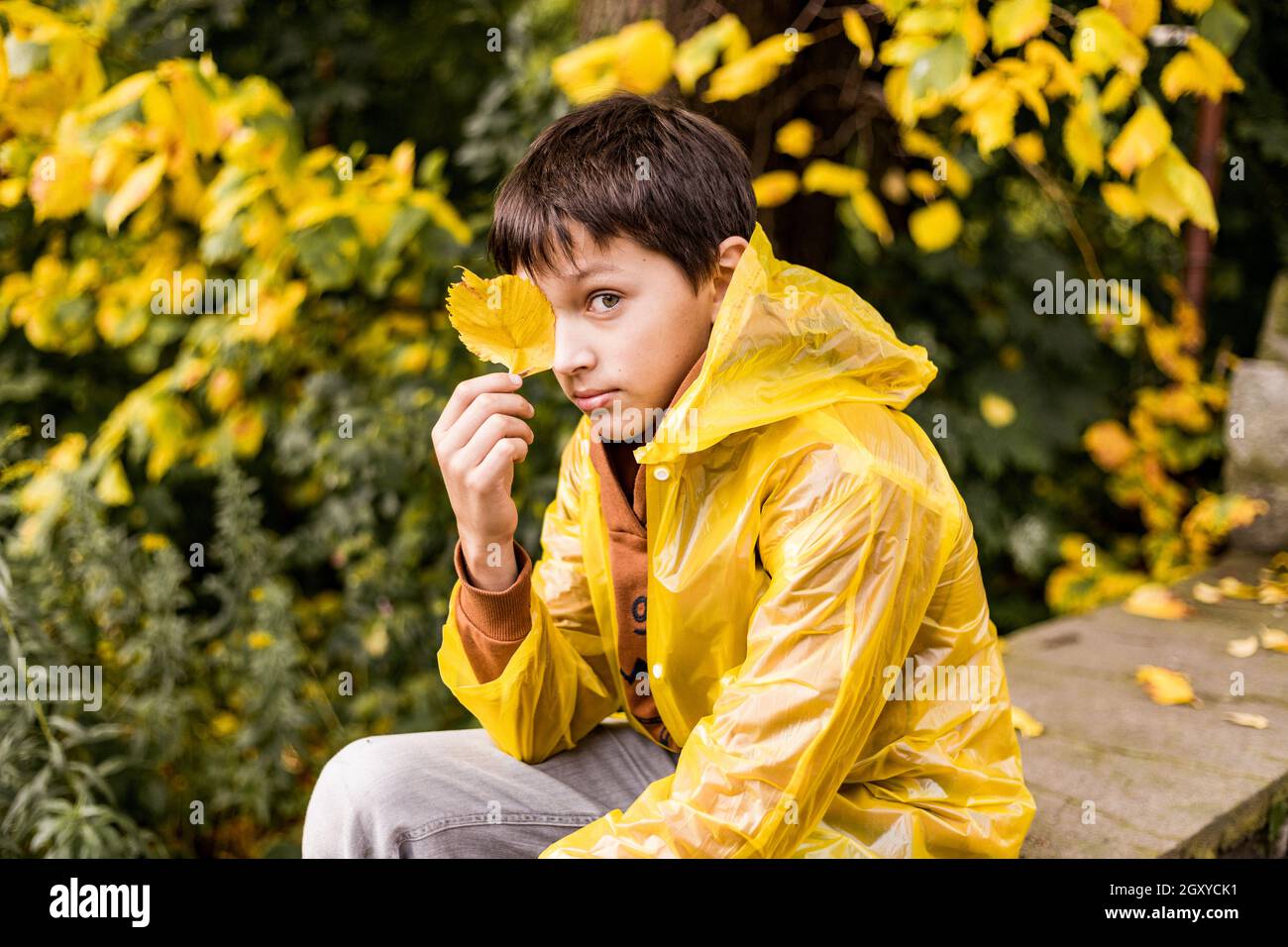 Photo of teenage boy in yellow raincoat among autumn leaves, closes one ...