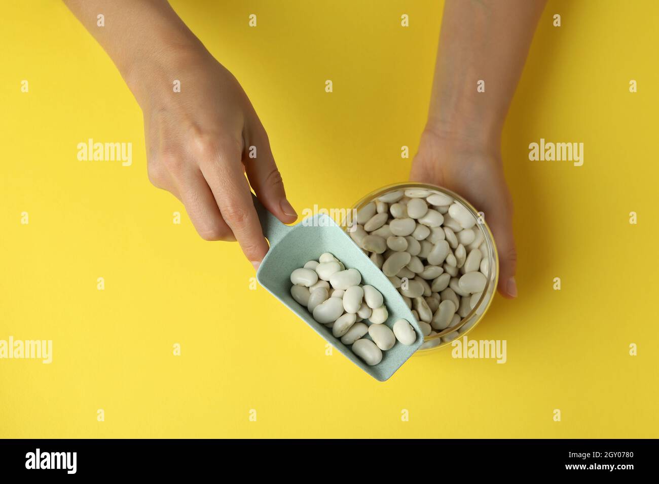Female hands hold jar and scoop with white beans on yellow background. Stock Photo