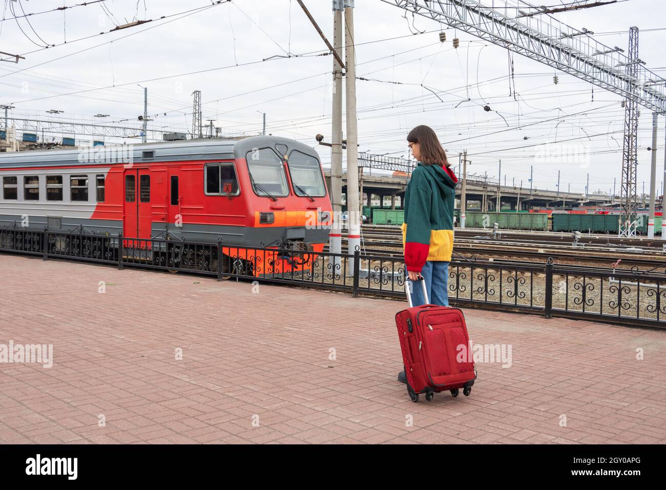 Caucasian girl student in bright clothes with a suitcase going to the electric train at the station. Girl with a suitcase at the train station. A trip Stock Photo