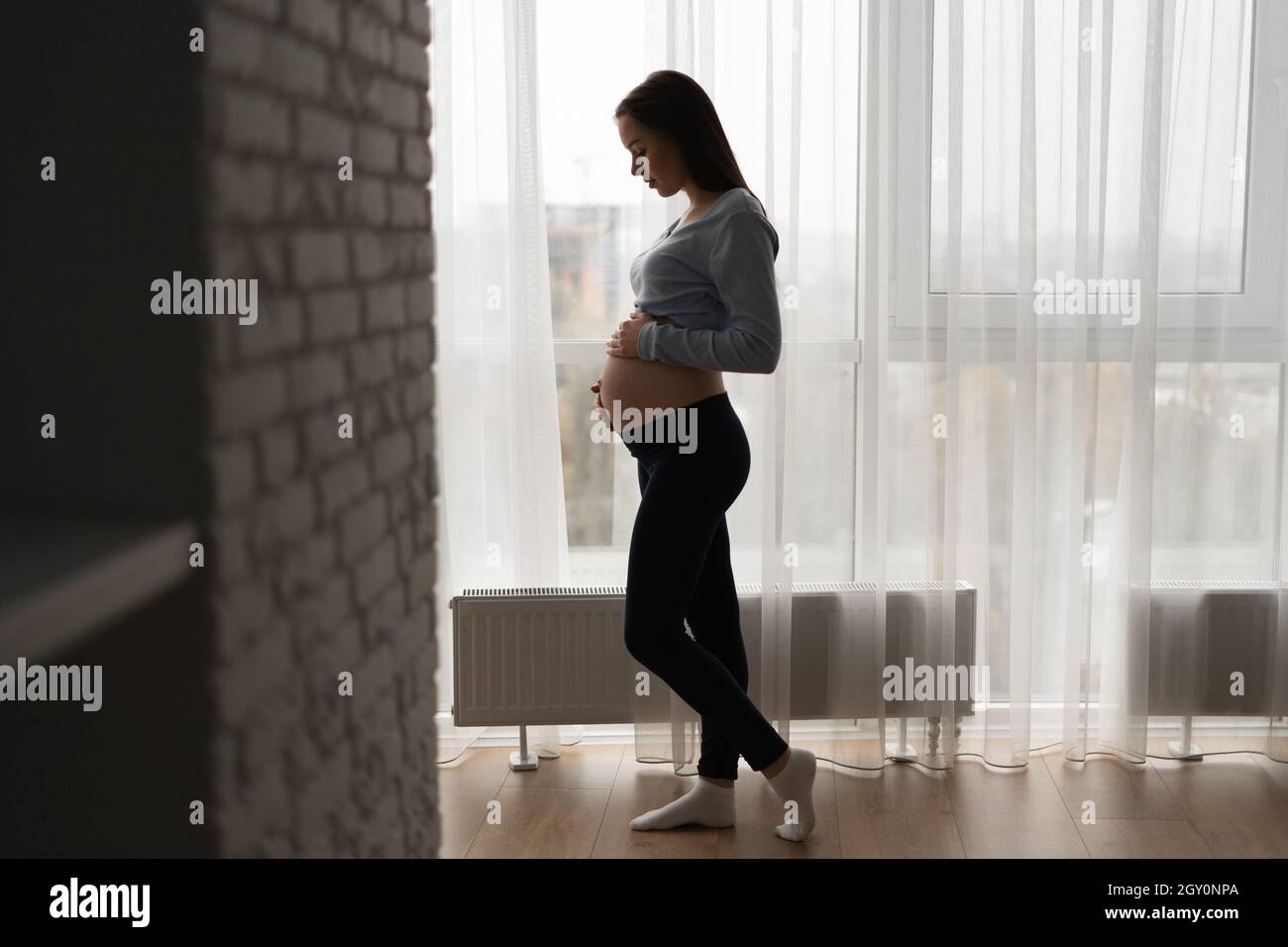 silhouette of a pregnant woman standing near a large window and stroking her big belly. Stock Photo