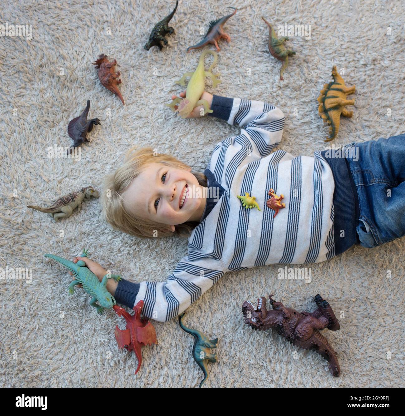 cute boy lies on light carpet on his back. There are many dinosaur figures spread out around it. little scientist, children's hobby - prehistoric anim Stock Photo
