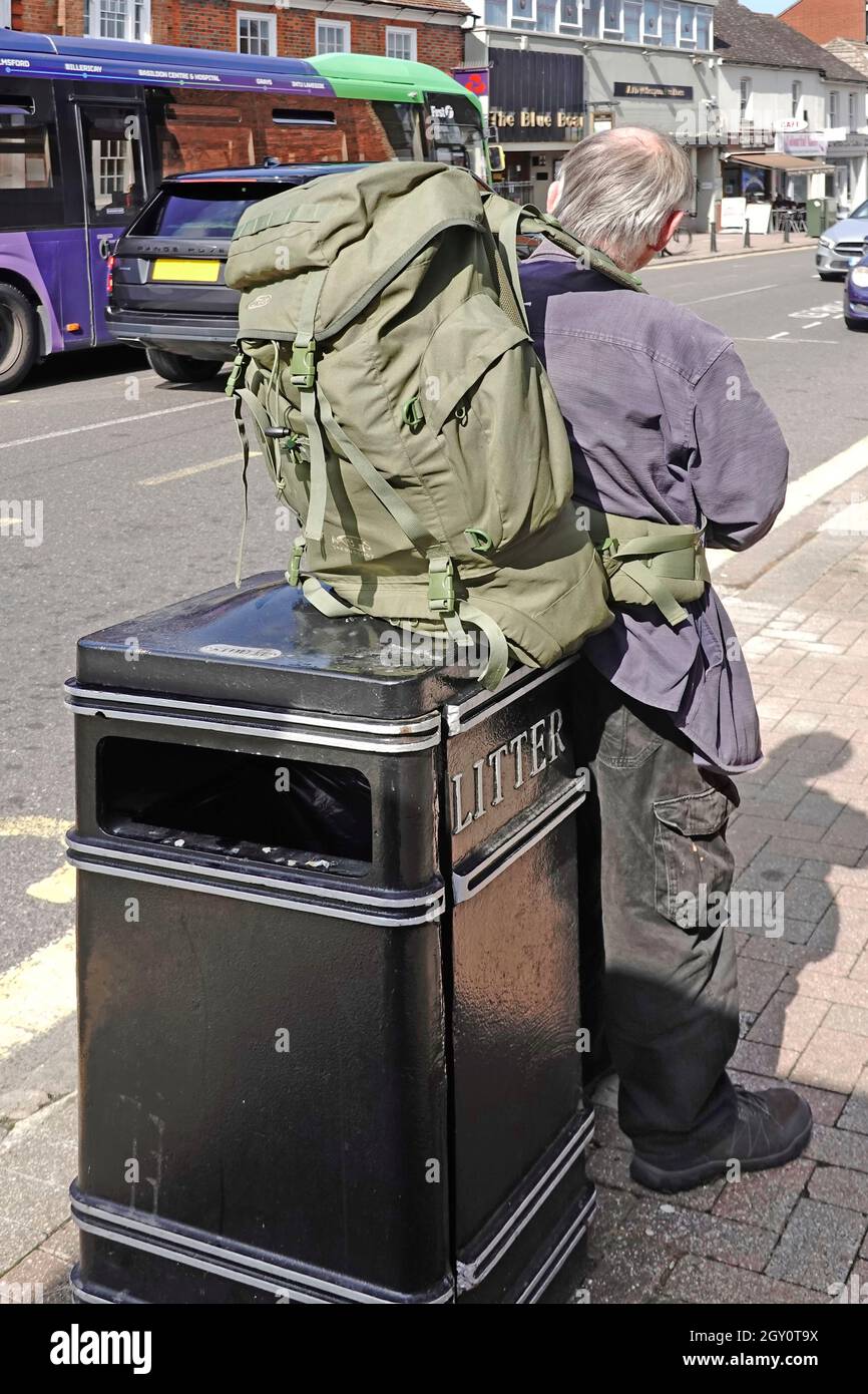 Town centre street scene grey haired man resting large heavy backpack on top of street furniture litter bin to take the weight off his feet England UK Stock Photo
