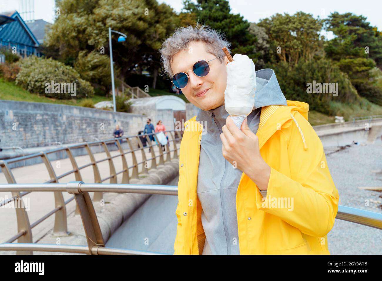 Excited caucasian young male tourist in dark sunglasses and yellow raincoat eating huge ice cream in a waffle cone during a walk near seaside. Positiv Stock Photo