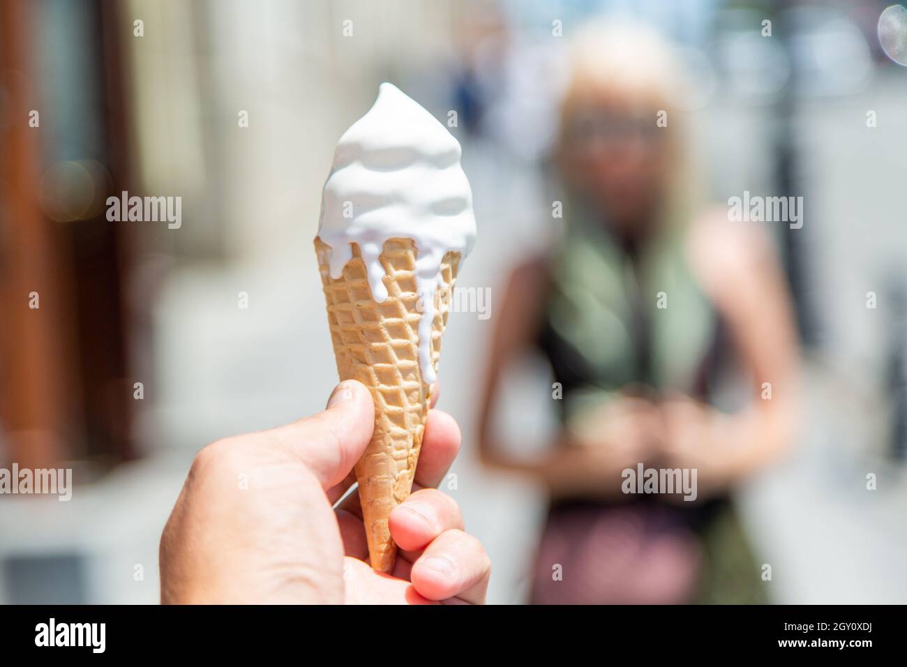 https://c8.alamy.com/comp/2GY0XDJ/in-the-foreground-a-mans-hand-holds-a-waffle-cone-with-white-ice-cream-which-melts-and-drips-down-it-2GY0XDJ.jpg