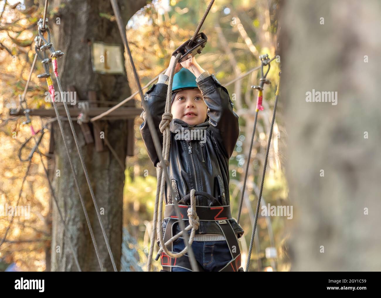 Climbing adventure park - little boy walking a track high up in the trees secured by safety equipment. Brave young child in safety harness climb high on tree tops Stock Photo