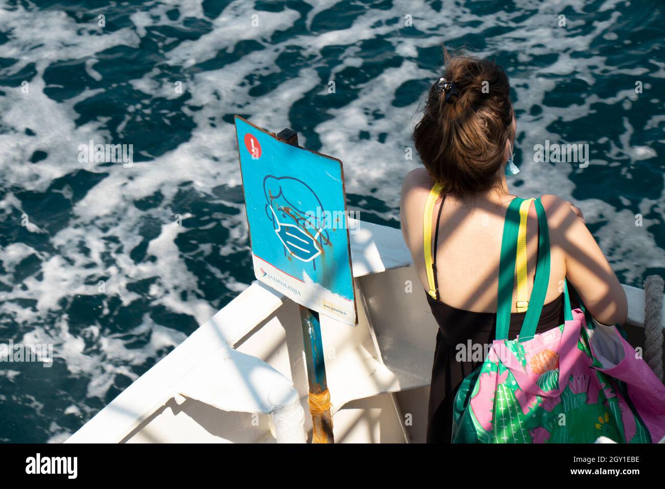 Vodice, Croatia - August 25, 2021: Wearing covid-19 face mask sign and one young woman passenger looking at sea water from a moving ship, high angle v Stock Photo