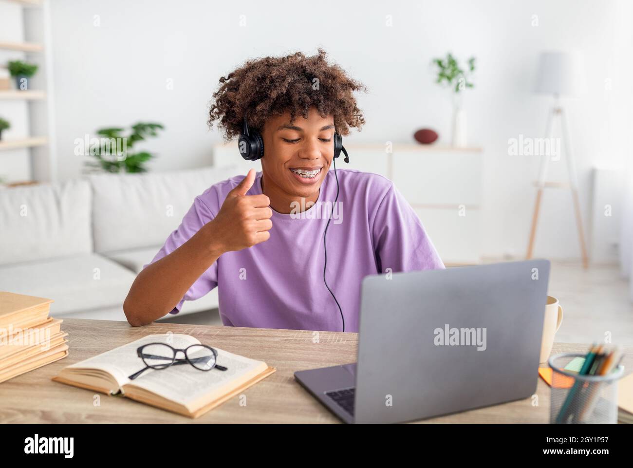 Funky black teen guy wearing headphones, having online class on laptop, showing thumb up at webcam indoors Stock Photo
