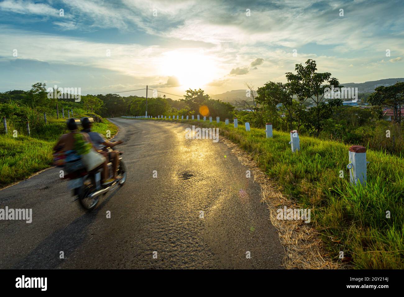 This is a couple traveling in a motocycle at the sunset in a lonely road at the counry side. The golden rays of the sun are reflected in the alphalt w Stock Photo