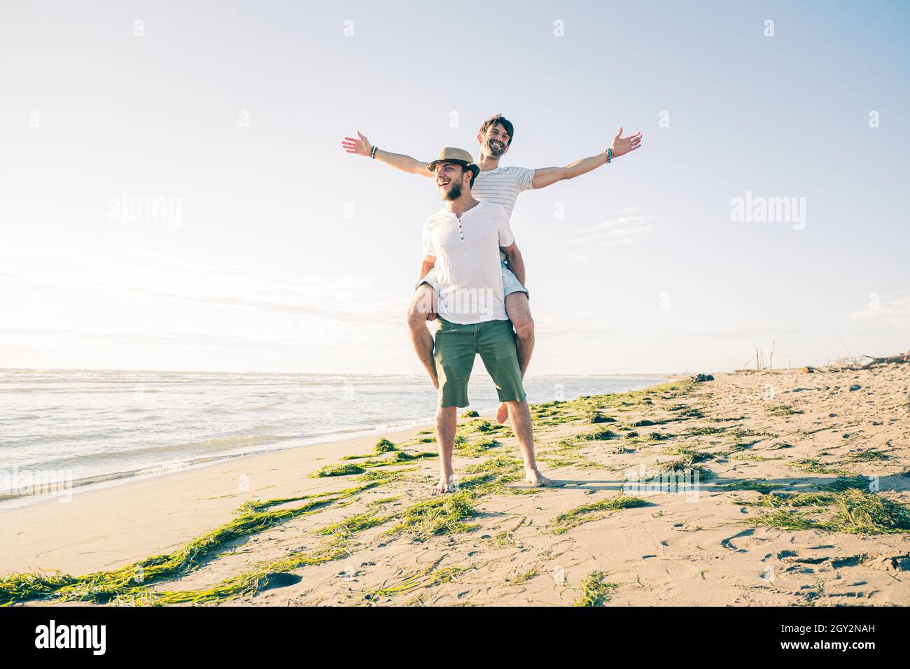 Two boy friends laughing and smiling and having fun at the beach -  Joyful guy piggybacking on young boyfriend Stock Photo