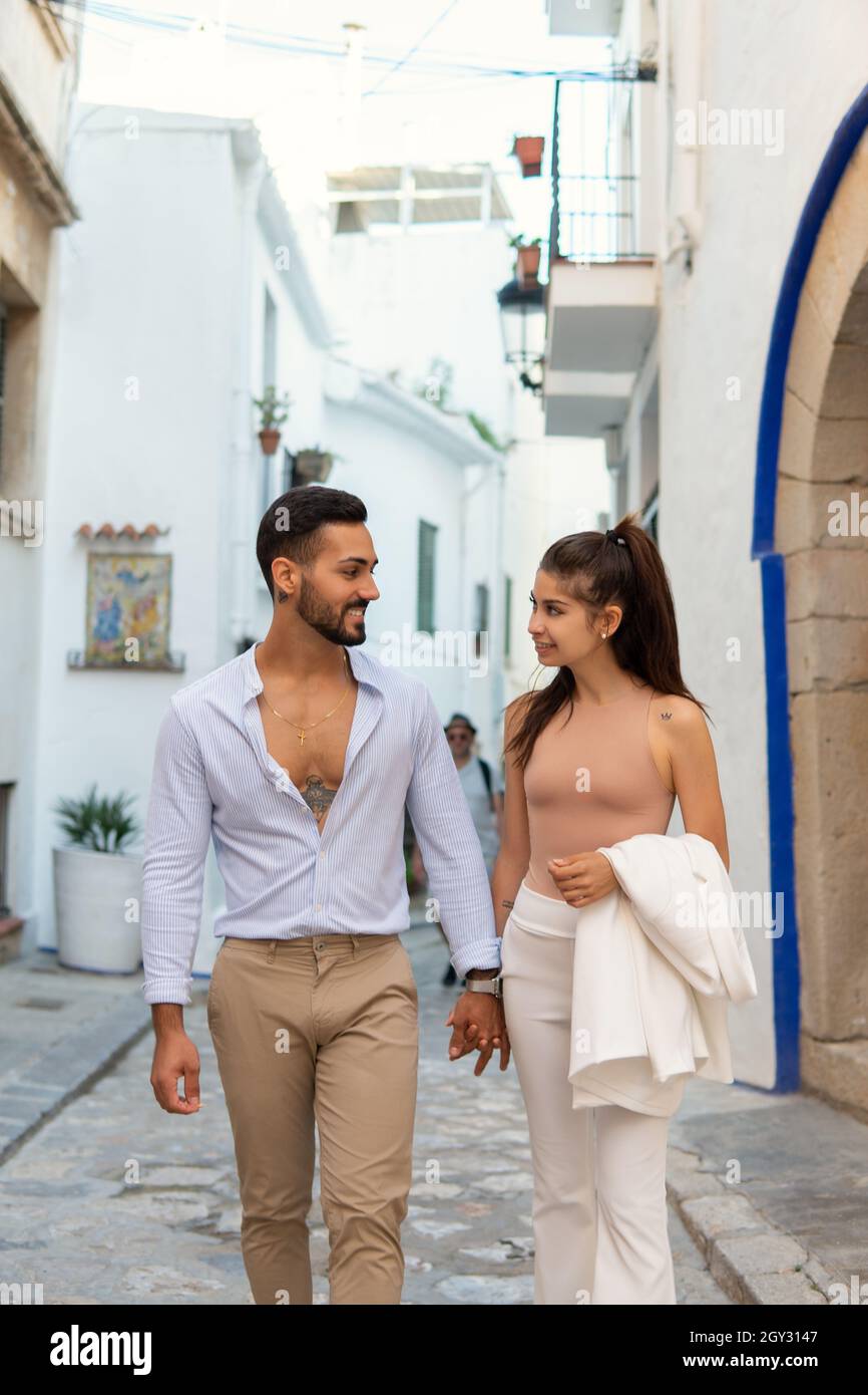 Trendy Hispanic couple in love holding hands and walking along old narrow street while looking at each other and smiling Stock Photo