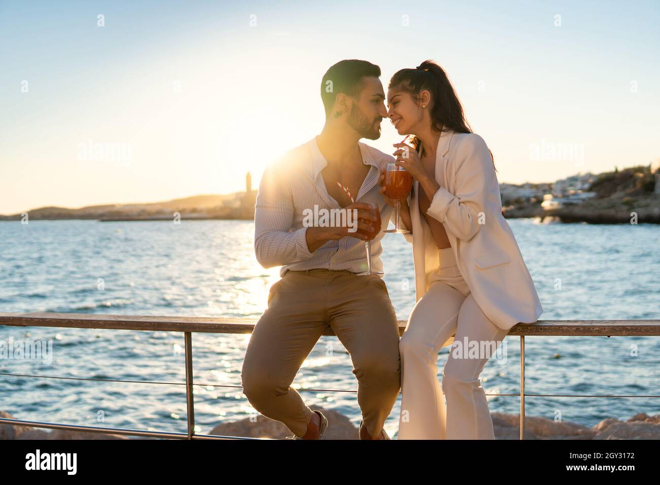 Content ethnic couple in love sitting close to each other on embankment against sea and drinking cocktails while looking at each other in summer evening Stock Photo