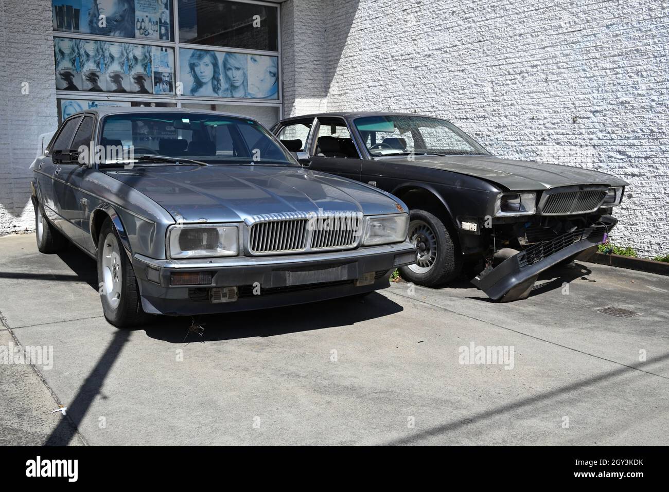 Two wrecked cars sitting outside a shop. Both are Jaguar XJ (XJ40) models, though one appears to be a Daimler-badged Double Six variant. Stock Photo
