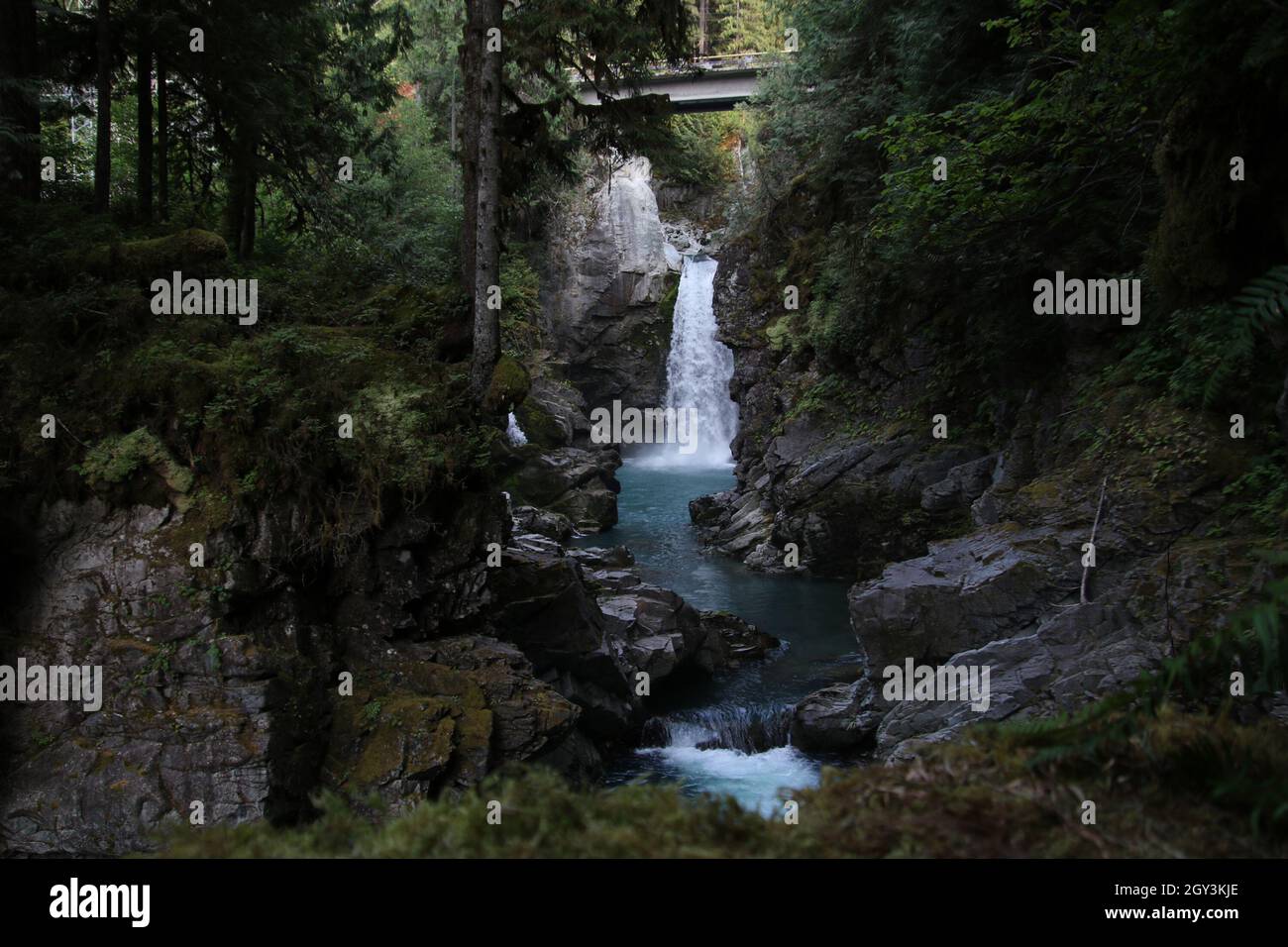 A waterfall flowing underneath a bridge flowing into a pool.  The river then flows between two rock walls. Stock Photo