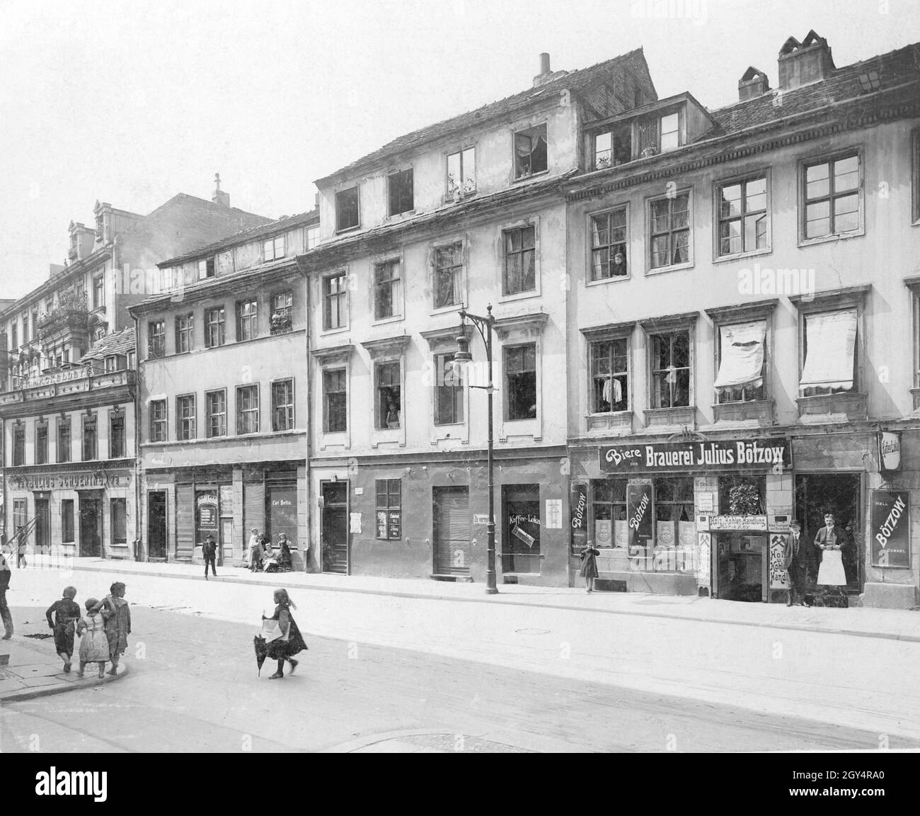 'Children run barefoot across a sidewalk in Neue Roßstraße, entrance Wallstraße in Berlin. On the right an innkeeper stands in front of his pub with beers of the Bötzow brewery. In the cellar there is a ''Holz u. Kohlen-Handlung'' of Marie Roy, who sells ''fishing worms'' and offers ''furniture transport and packing'' at the same time. To the left is a pub for rent, next to it is a ''Besohl-Anstalt'' and the shop of Julius Schoening. Photo undated, taken around 1905. [automated translation]' Stock Photo