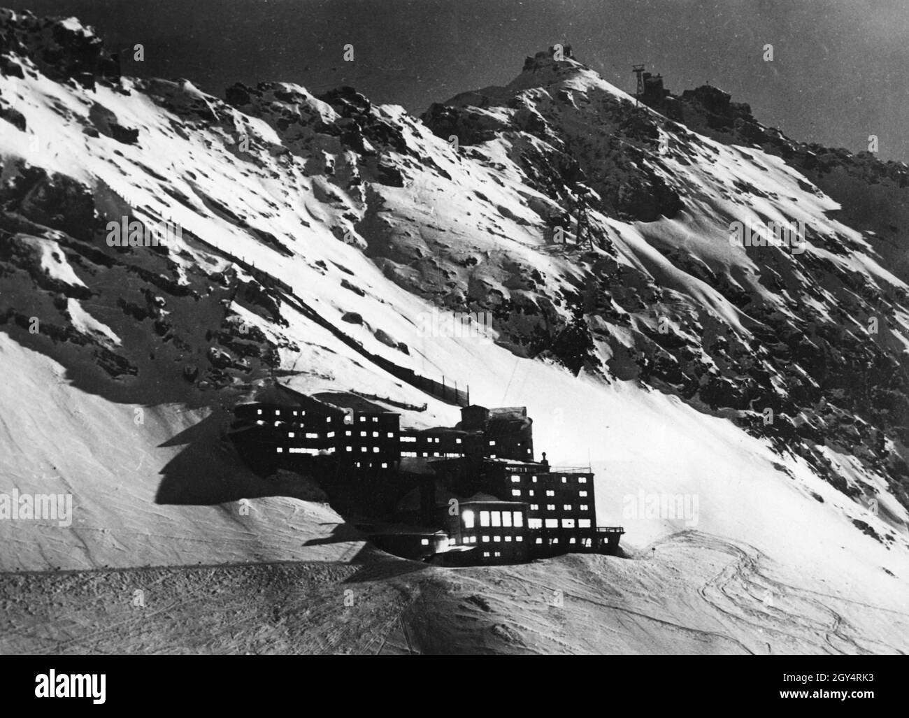 The Schneefernerhaus below the Zugspitze peak is situated with brightly lit windows in the middle of the wild snow and rock landscape, which is illuminated by pale moonlight at night. The house is connected to the summit by cable car. Ski tracks lead downhill from the house. The photograph was taken on 2 November 1934 [automated translation] Stock Photo
