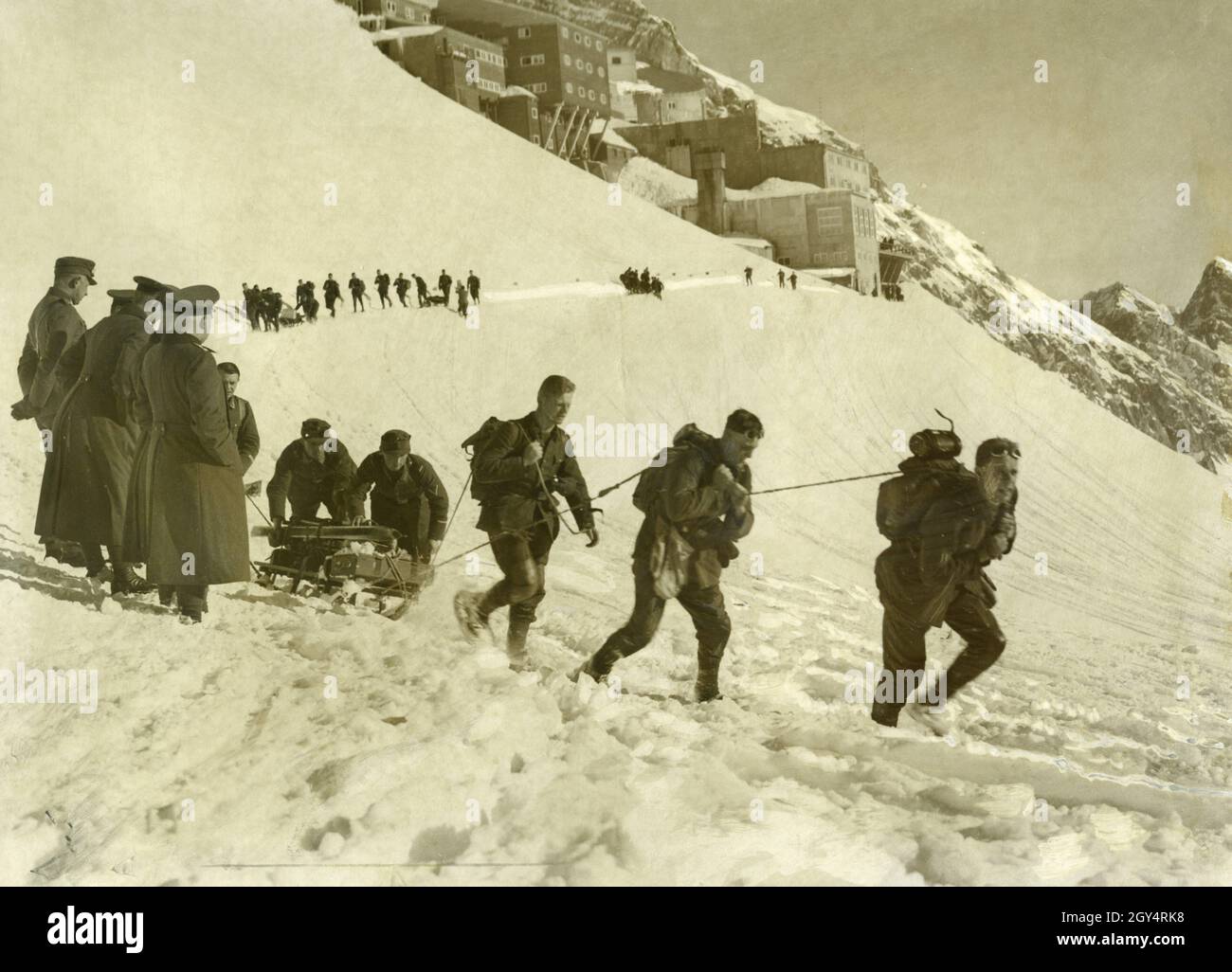 In 1934, exercises of the mountain troops took place on the Zugspitzplatt. In the picture, a column has set off from the Schneefernerhaus below the Zugspitz peak and is now marching past its officers up the snowy slopes. Several soldiers at a time are pulling and pushing one of the sledges, which carry the necessary equipment such as skis. The soldiers are wearing snowshoes. [automated translation] Stock Photo