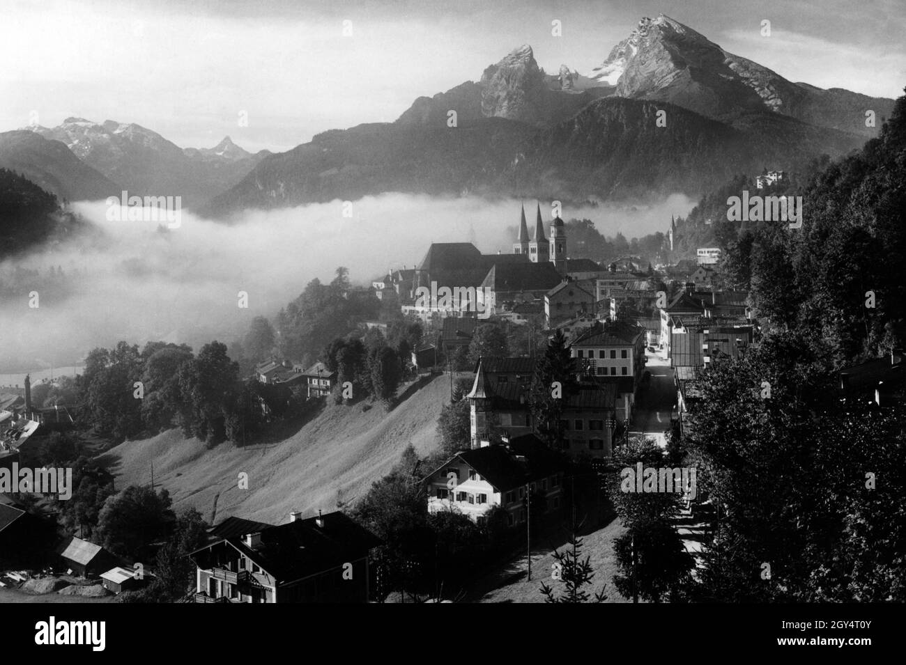 'The photograph shows Berchtesgaden on a summer morning in 1936 as seen from Locksteinstraße. In the foreground is the ''Villa Rössler'' (with corner tower), behind it the churches and steeples of St. Andreas, the collegiate church (behind) and the Christuskirche (right, far back). Out of the morning mist, which lies over the river valley and the Königssee, the alpine chain with its mountain peaks stands out (from left to right): Funtenseetauern, Schönfeldspitze, Watzmann, Grünstein (directly in front). [automated translation]' Stock Photo