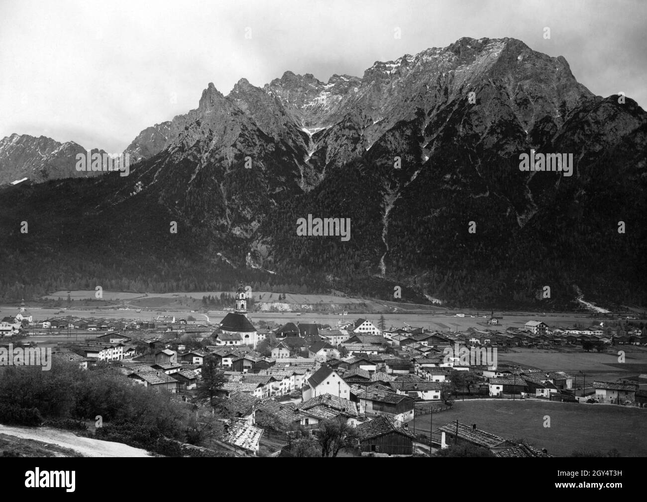 This photograph, taken around 1910, shows Mittenwald with the church of St. Peter and Paul in the centre and St. Nikolaus on the left. Behind the village rises the Northern Karwendel range with Karwendelkopf, Karwendelspitze and Linderspitze. Photo from the art publishing house of Max Stuffler from Munich. [automated translation] Stock Photo