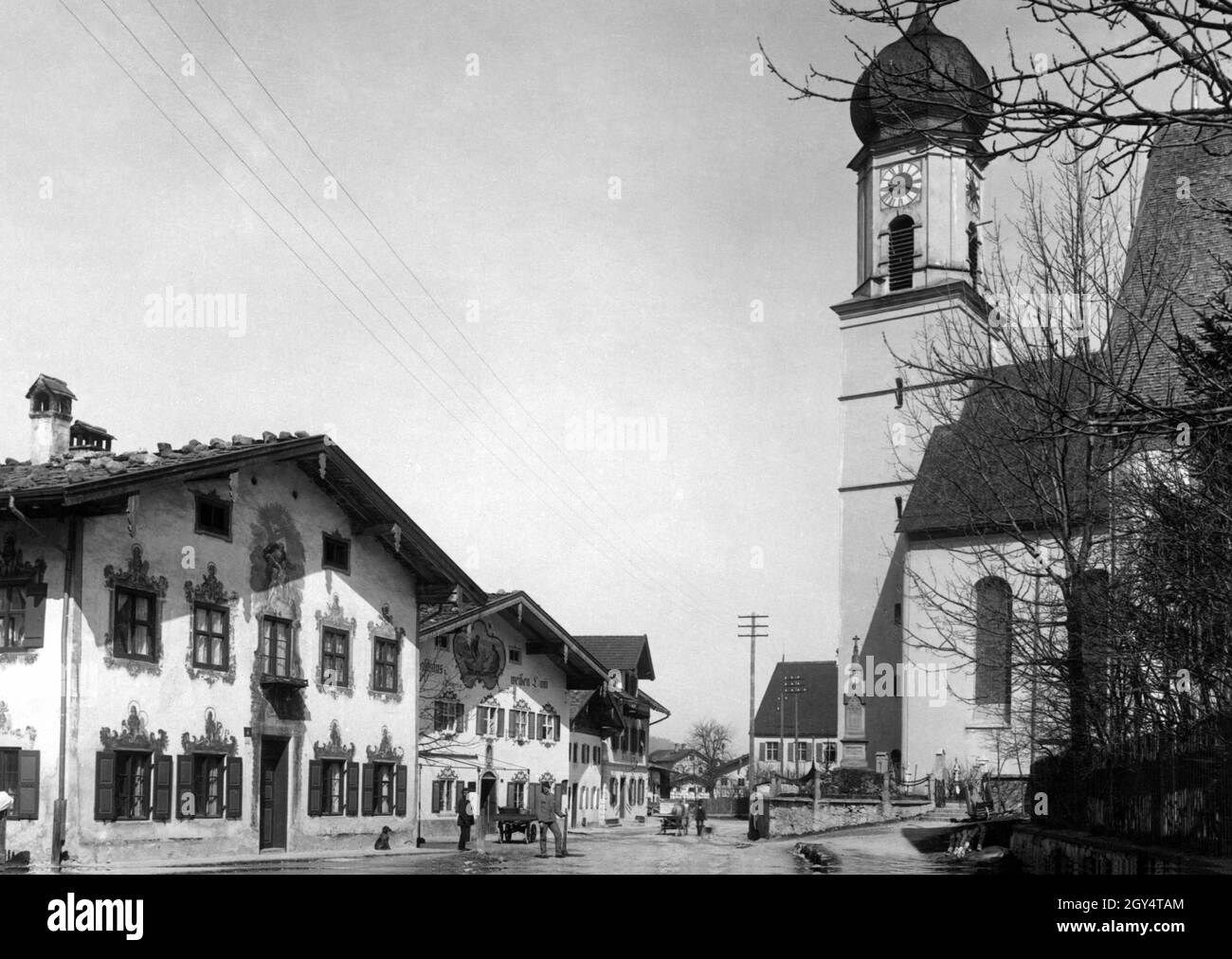 'Two workers stand in the sunshine on Ettaler Straße in Oberammergau around 1930. On the right is the parish church of St. Peter and Paul, on the left the houses decorated with frescoes, including the ''Gasthaus zum weißen Lamm'' (so-called Dedlerhaus), and the Kölblhaus (far left). [automated translation]' Stock Photo