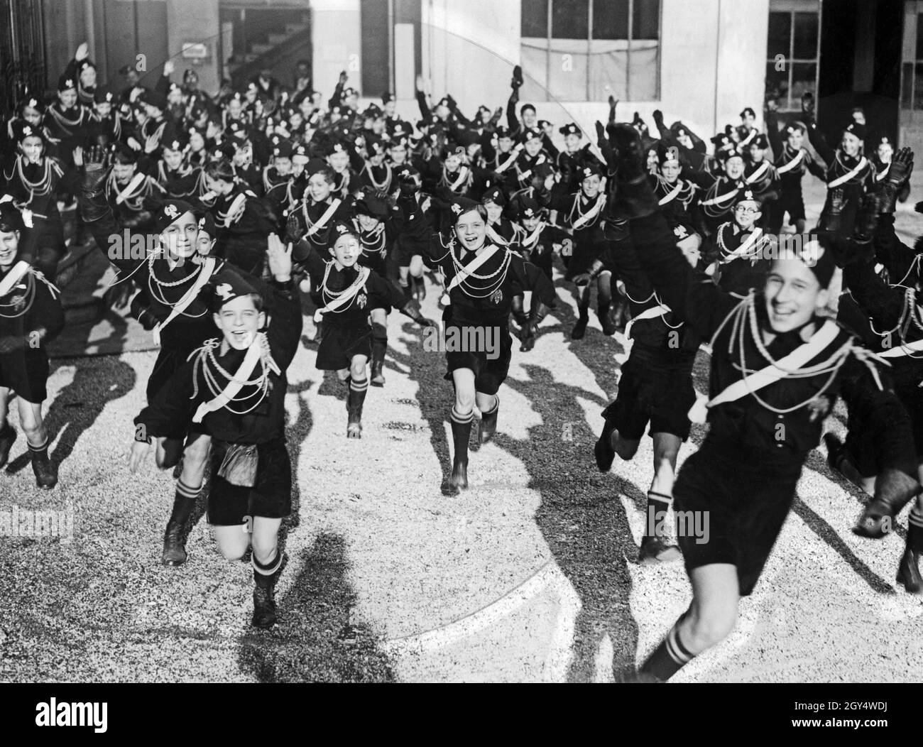 Dozens of children and young people from the Opera Nazionale Balilla run disorderly across a courtyard, cheering as they go. Some raise their arms in a saluto romano. The photograph was taken in September 1928. [automated translation] Stock Photo