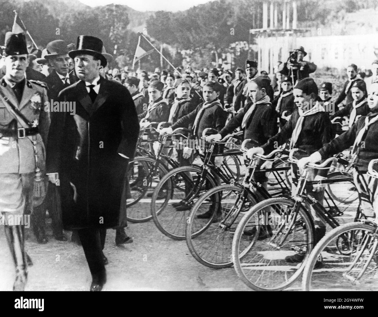 Benito Mussolini (left, in civilian clothes) strides along the front of a bicycle division with officers and guests. The boys belong to the fascist youth organization Opera Nazionale Balilla. Retouched photograph, taken around 1930. [automated translation] Stock Photo