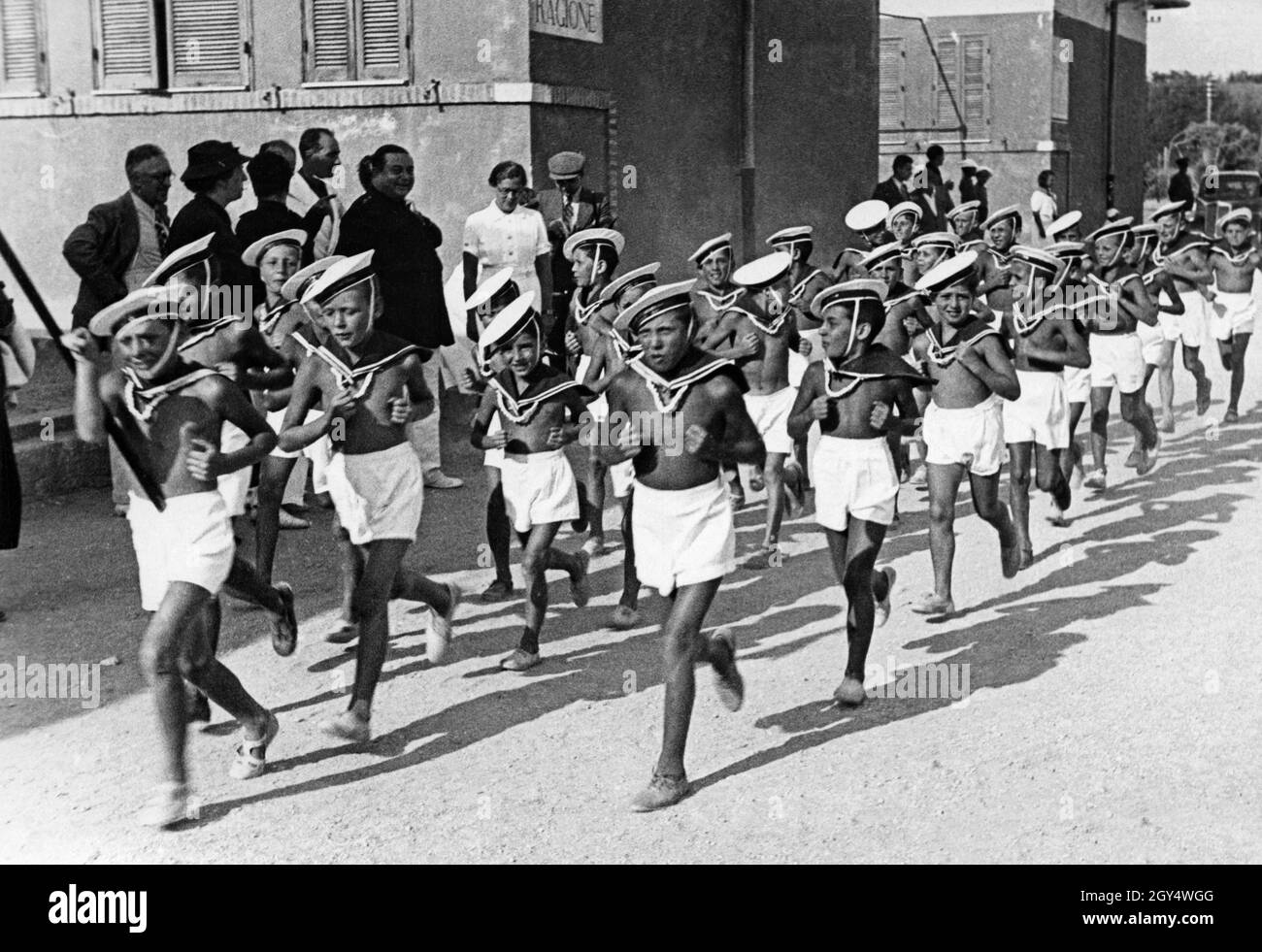 'A group of children and teenagers, dressed only in shorts and berets, walk through a street of the Colonia marina di Anzio in Italy in the summer of 1937. They belong to the Marinaretti, the marine training division within the fascist youth organization Opera Nazionale Balilla. Sports exercises such as endurance running were used by the Italian Fascists for the ''physical training'' of youth. [automated translation]' Stock Photo