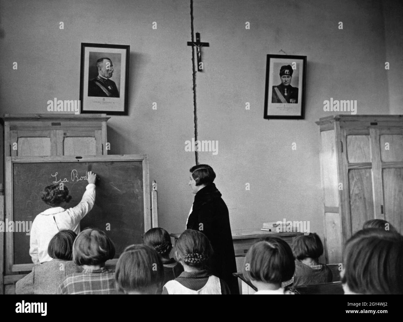 A student writes on the blackboard with chalk in front of her class and the teacher. On the wall hang a picture of the King of Italy, Victor Emmanuel III. (left), a crucifix and a picture of the Italian dictator, Benito Mussolini (right). The girls' class is taught at the newly built school of Latina (then Littorio). Undated photograph, taken around 1935. [automated translation] Stock Photo