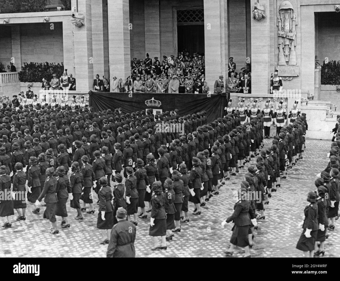 Adolf Hitler visited Italy, allied with Nazi Germany, at the beginning of May 1938. On Friday, May 6, 1938, a parade with 50000 soldiers and members of fascist organizations took place on the Via Trionfale (today Via di San Gregorio) in Rome. The picture shows the female fascist legion parading in rows of 24, passing the tribune. On the tribune are (from left to right): Benito Mussolini, Adolf Hitler, King Victor Emmanuel III of Italy and his wife, Elena of Montenegro. Behind them line up other greats of both fascist states. [automated translation] Stock Photo