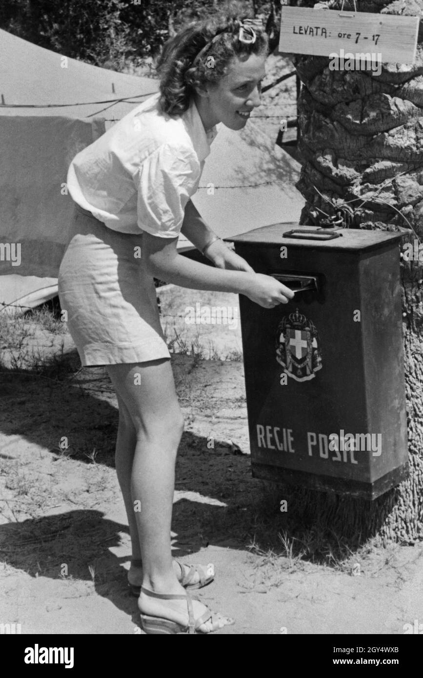'A young Italian fascist throws a letter into the mailbox of the ''Regie Poste'' (Royal Post Office), which has been attached to a palm tree. It is emptied daily at 7 a.m. and at 5 p.m. (''Levata: ore 7 - 17''). The woman is in a colonial camp in the Italian colony of Libya. Undated photograph, probably taken in the late 1930s. [automated translation]' Stock Photo