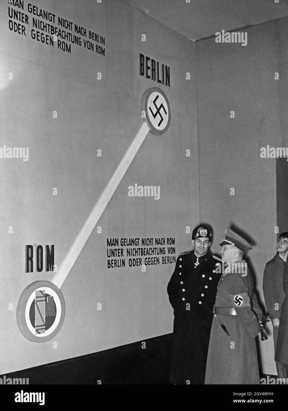 'On November 6, 1937, the day Italy joined the Anti-Comintern Pact, an ''Anti-Bolshevik Exhibition'' entitled ''Bolshevism without a Mask'' was opened in the Reichstag in Berlin. The picture shows a room of the exhibition in which the Berlin-Rome axis was visually represented. Looking at the line between the two capitals of Rome (with the fascist symbol Fascis) and Berlin (with the Nazi symbol Swastika) is the head of the Italian section of the exhibition, General Alessandro Melchiori (left) and Gaupropaganda director of Berlin, Werner Wächter (right). On the wall there are two versions of a Stock Photo
