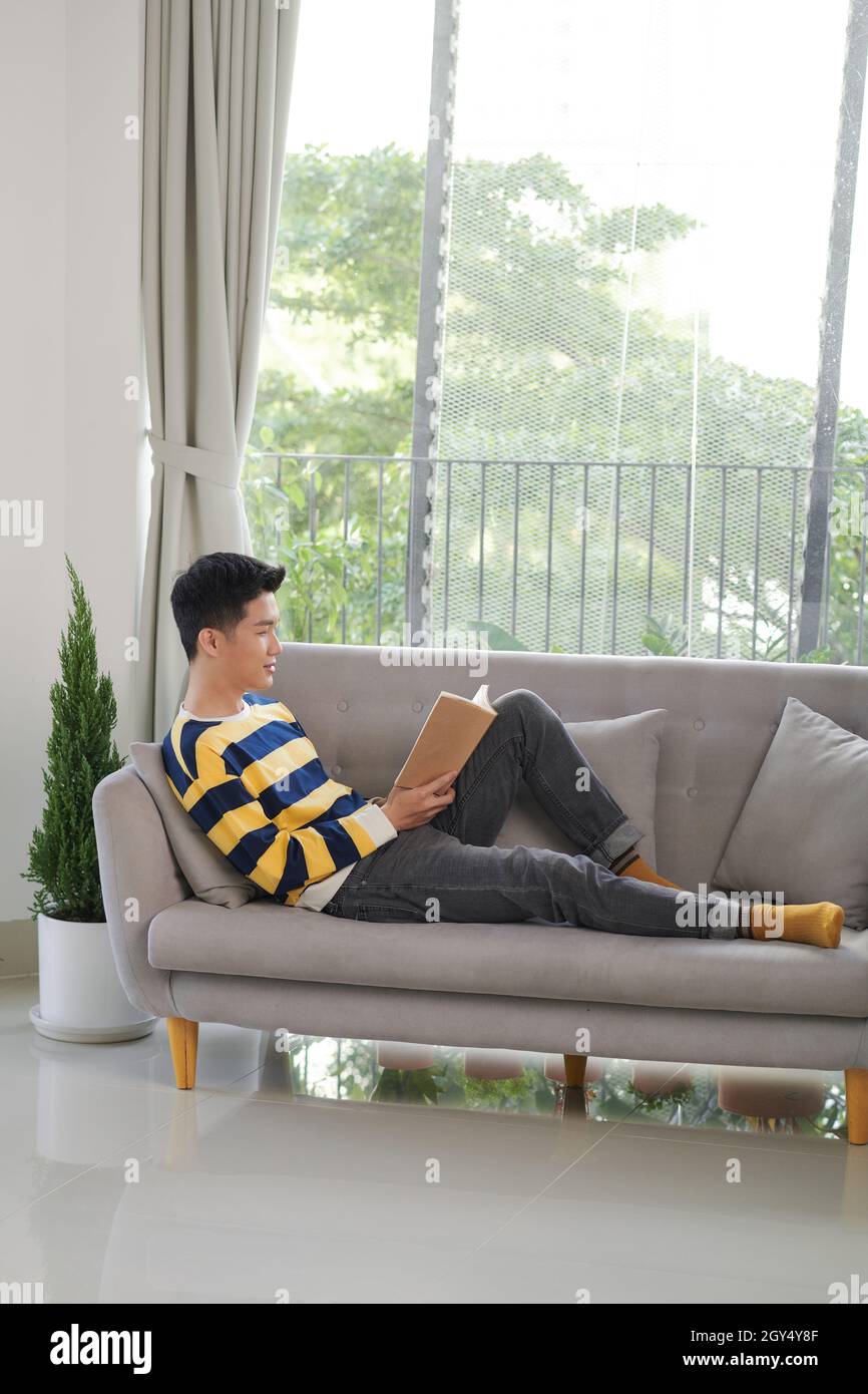 Handsome man in casual wear smiling lying on sofa reading handheld book. Stock Photo