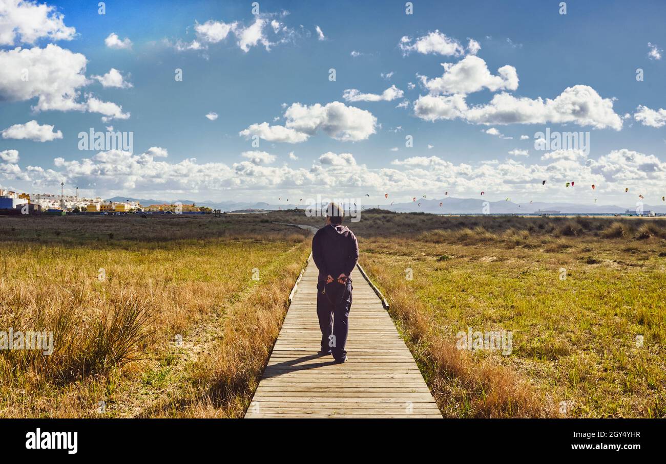 Man walking along a wooden boardwalk through grassland towards distant kite surfers in a sunny cloudy sky with whitewashed buildings to the side in a Stock Photo
