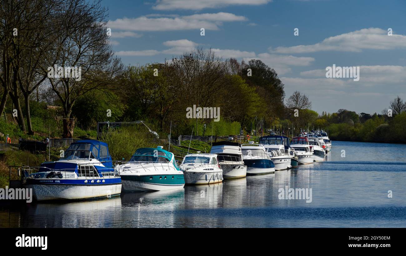Line of sunlit boats (leisure craft) moored on River Ouse at riverbank moorings on sunny summer day - Fulford, York, North Yorkshire, England, UK. Stock Photo