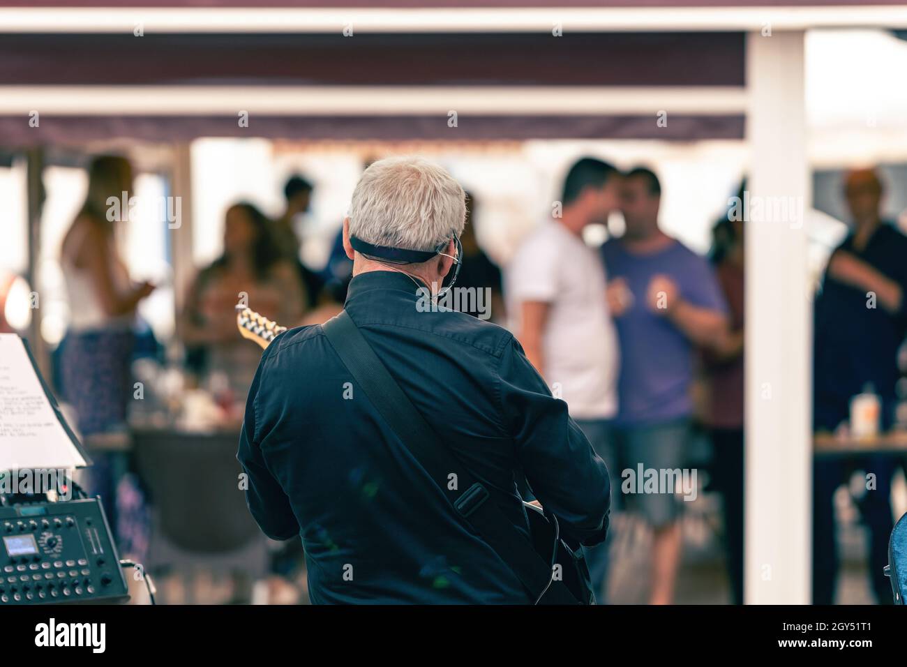 Back of an old man playing the electric guitar on a terrace with public Stock Photo