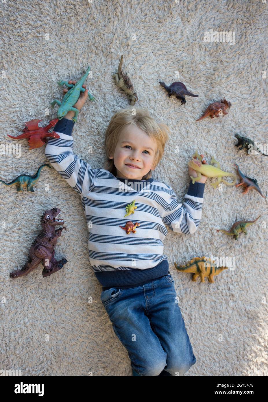 lot of dinosaur figures are spread out around boy of 4-5 years old lying on light carpet on back. little scientist, child's hobby - prehistoric animal Stock Photo
