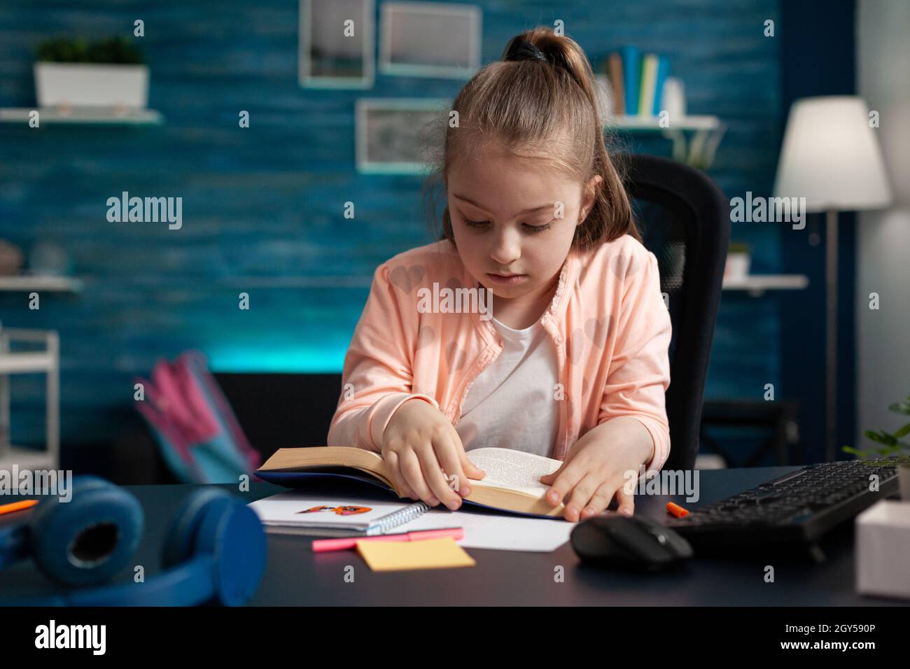 Schoolchild sitting at desk in living room holding school book reading educational literature story for online course during coronavirus quarantine. Reader child studying for academic exam Stock Photo