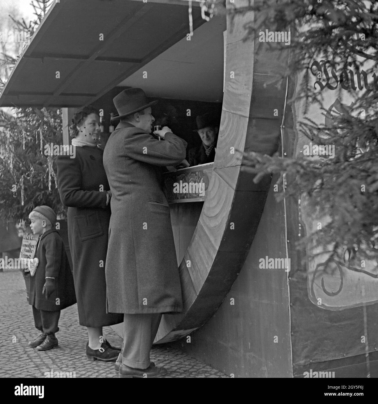 Ein kleiner Junge langweilt sich, während seine Eltern am Glühweinstand auf dem Weihnachtsmarkt stehen, Deutschland 1930er Jahre. A little boy is bored beyond belief while his parents having hot wine punch at the christmas market, Germany 1930s. Stock Photo