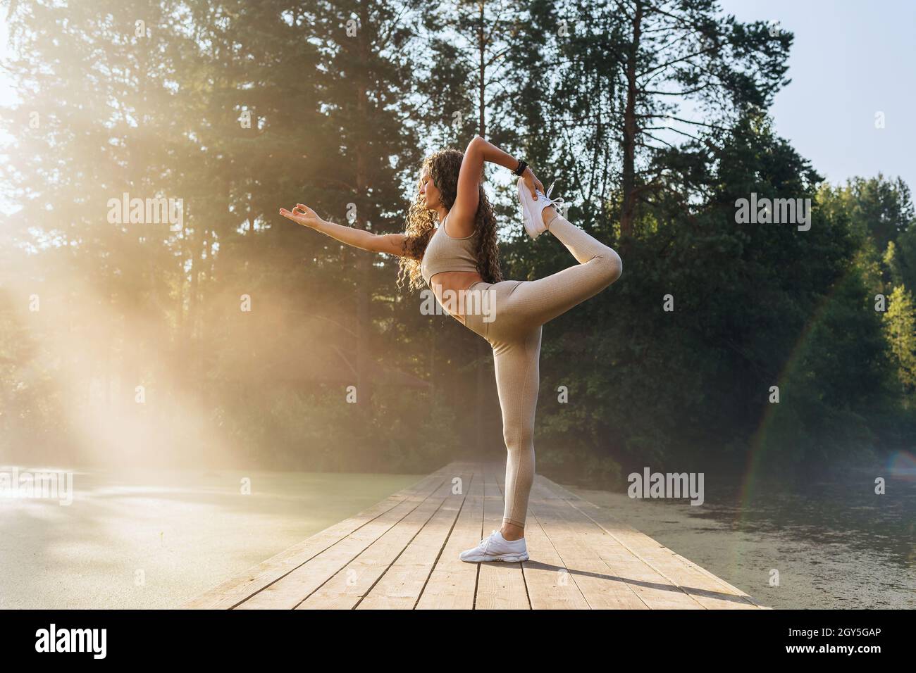A woman practicing yoga, performs the natarajasana exercise, the pose of the king of dancers with an outstretched arm, stands on a wooden bridge in a Stock Photo