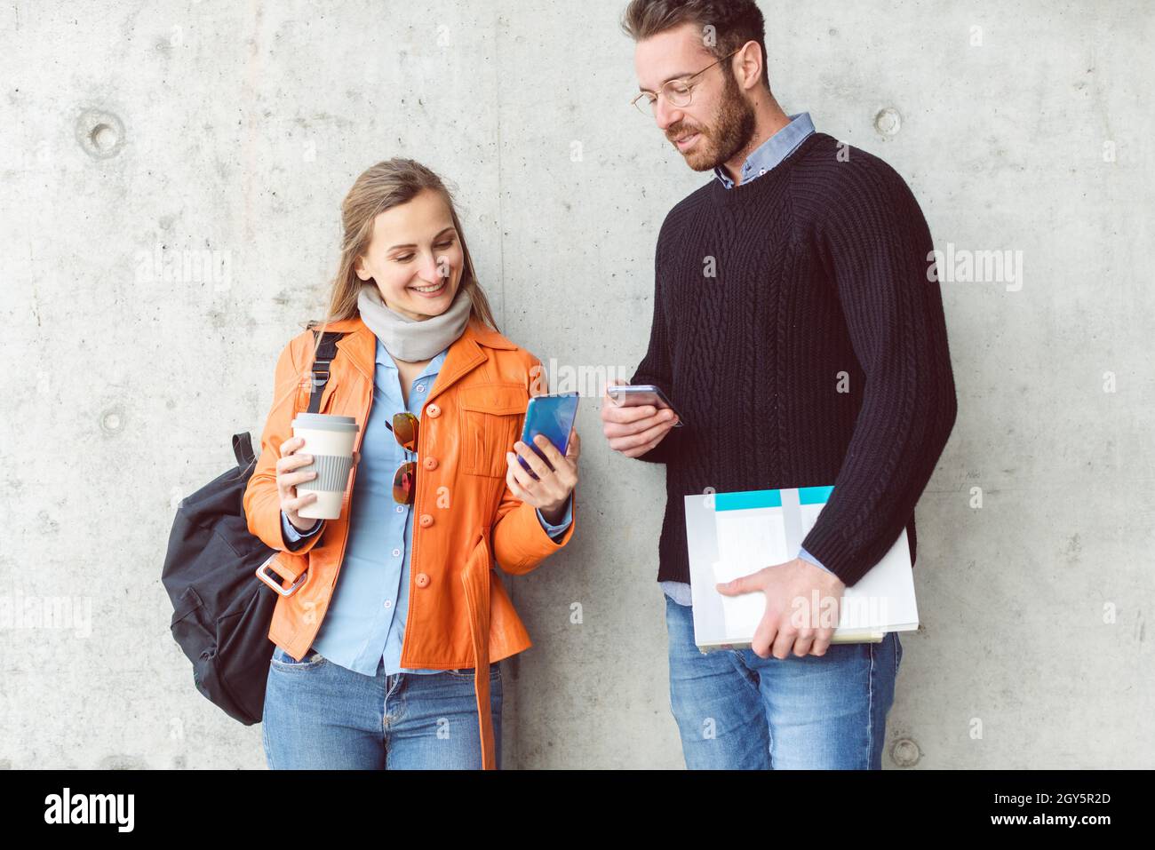 Two students with phone and books standing in front of concrete wall on campus Stock Photo