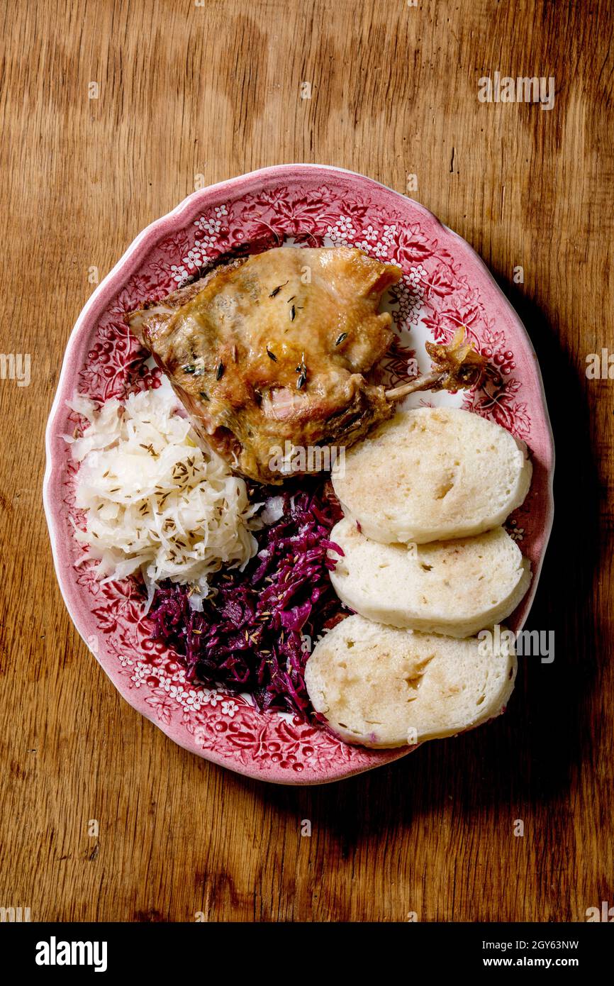 Baked duck legs with sliced boiled bread knedliks and sauerkraut in ceramic plate over brown wooden background. Traditional Czech, German and european Stock Photo
