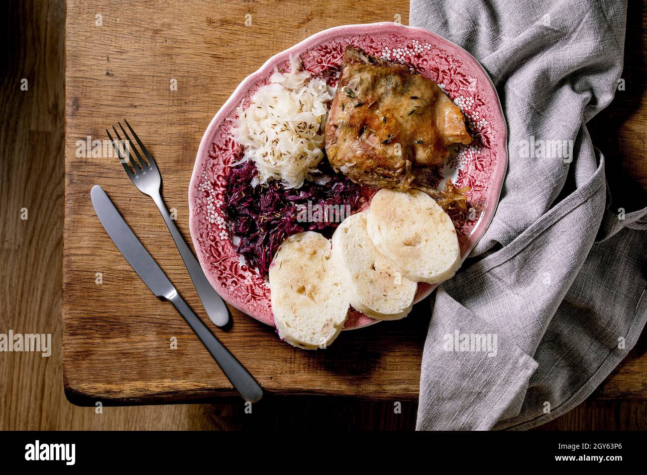 Baked duck legs with sliced boiled bread knedliks and sauerkraut in ceramic plate, decorated with napkin over brown wooden table. Traditional Czech, G Stock Photo