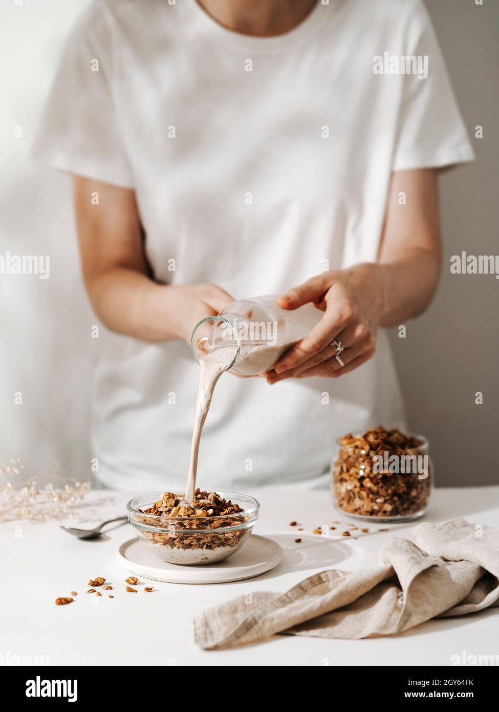 Unrecognizable woman pouring vegan non-dairy milk into glass bowl with gluten-free granola or muesli. Vertical. Modern light minimal pastel color pale Stock Photo