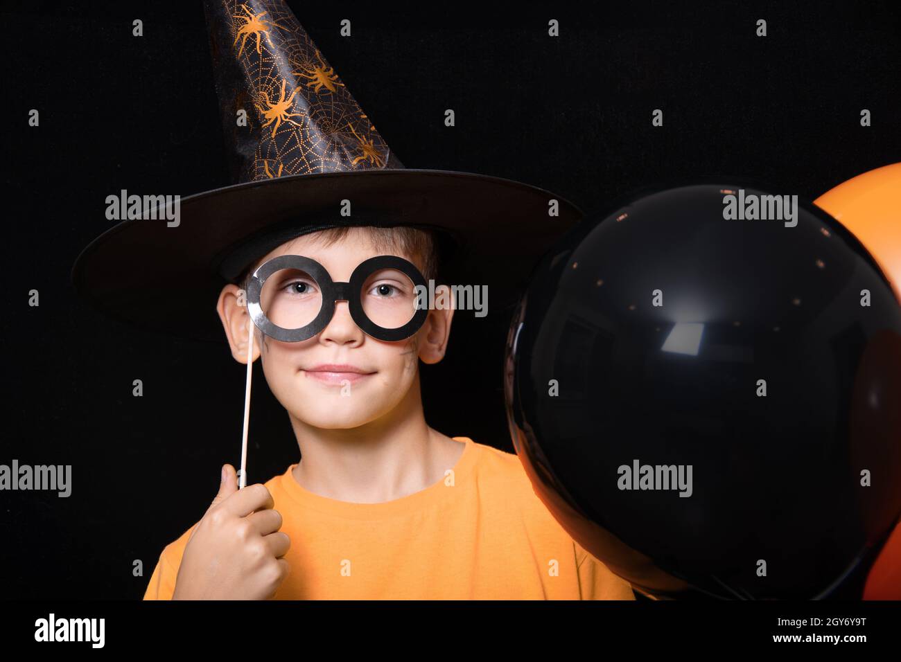 Halloween kids. A boy in a magician's hat and with a mask of black glasses holding orange and black balloons on a black background. Ready for the holi Stock Photo