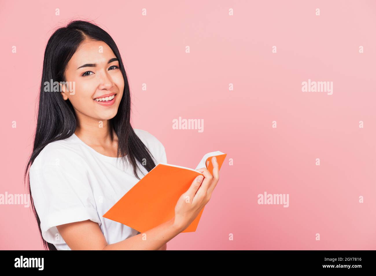 Portrait of happy Asian beautiful young woman confident smiling standing holding orange book open or diary for reading, studio shot isolated on pink b Stock Photo