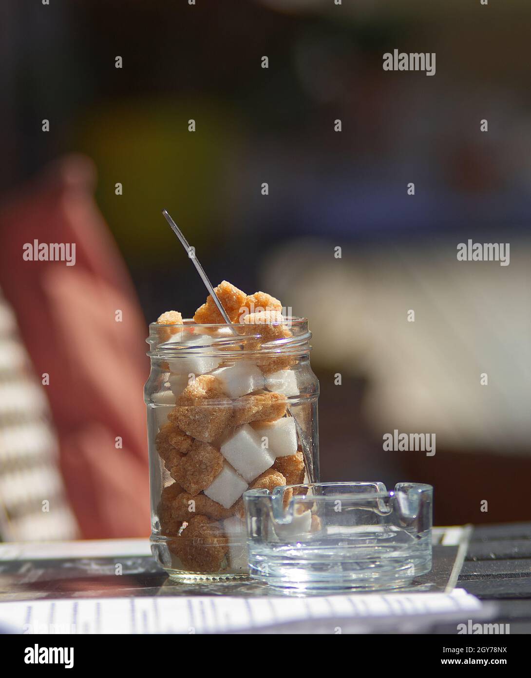 A jar of cube sugar, brown and white cube sugar in a jar on a coffee table in the street, with a small ashtray next to it. Blurred background. Stock Photo